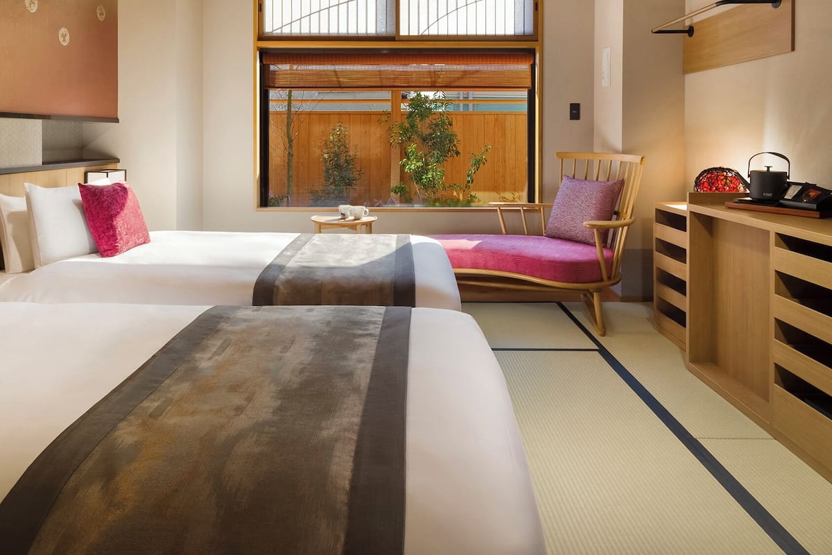 A hotel room with two beds, a wooden chair, a small table, and a view of the outside.