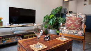 A living room with a television, two wine glasses on a wooden table, a floral armchair, and a potted plant.
