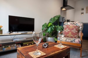 A living room with a television, two wine glasses on a wooden table, a floral armchair, and a potted plant.