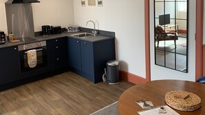 A modern kitchen with a black range hood, stainless steel appliances, and a dark blue island. There is a small trash can and a round wooden table with a woven placemat and a menu.