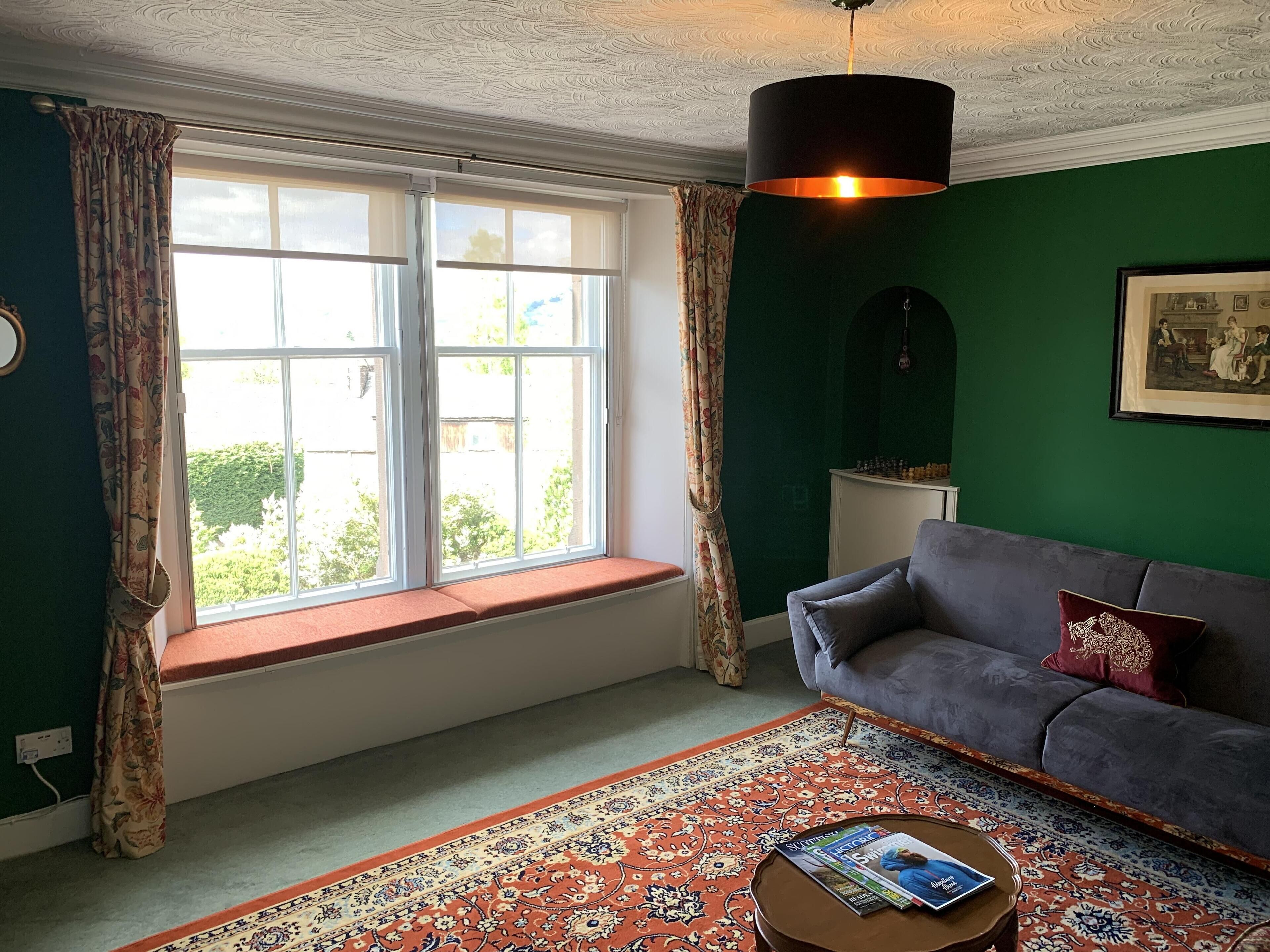 A room with a green wall, a grey sofa, a window seat, and a patterned rug.