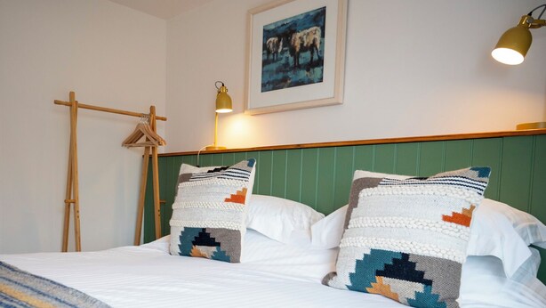 A neatly made bed with two pillows, a green headboard, a framed picture on the wall, and a hanging light fixture.