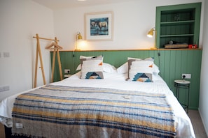 Een bed met een gestreepte deken, twee kussens, een lijst met een schilderij, een lamp en een groene plank met een radio.
