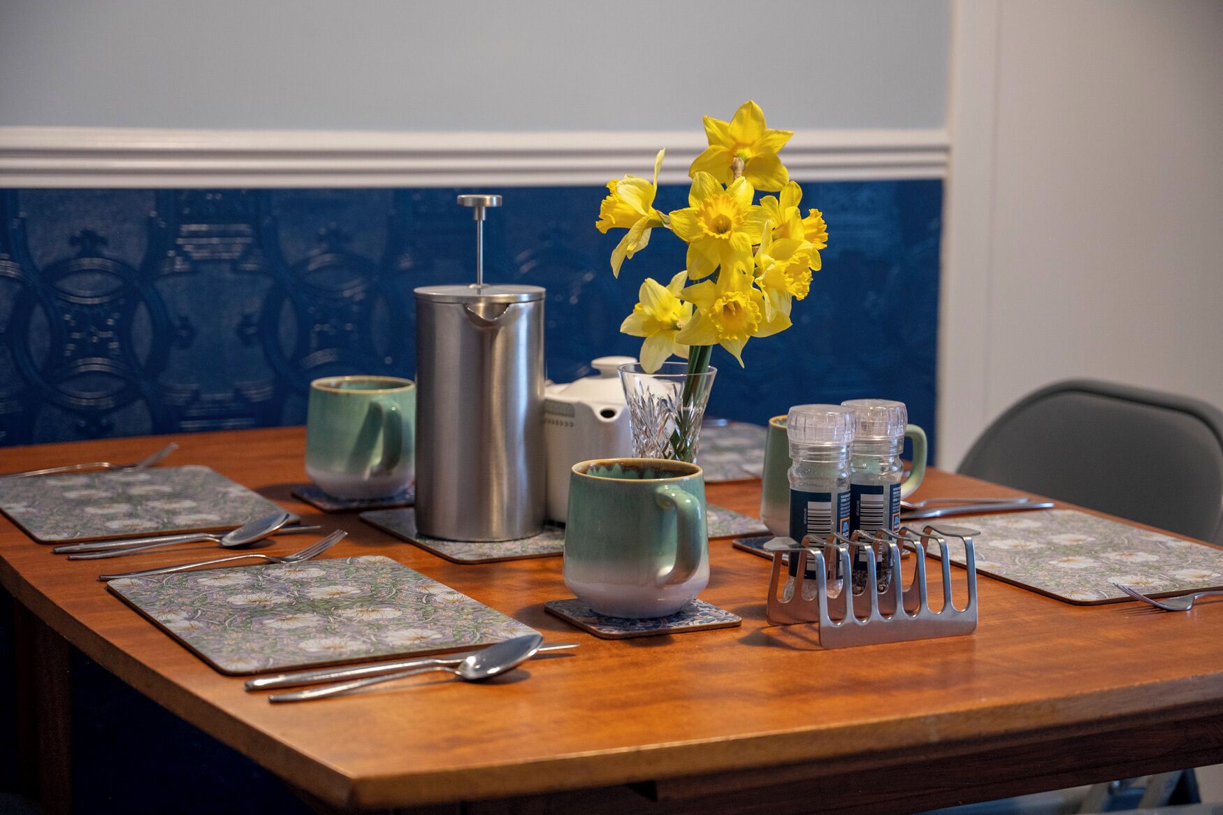 A dining table set with plates, cutlery, a coffee pot, a vase with yellow flowers, and a mug rack.