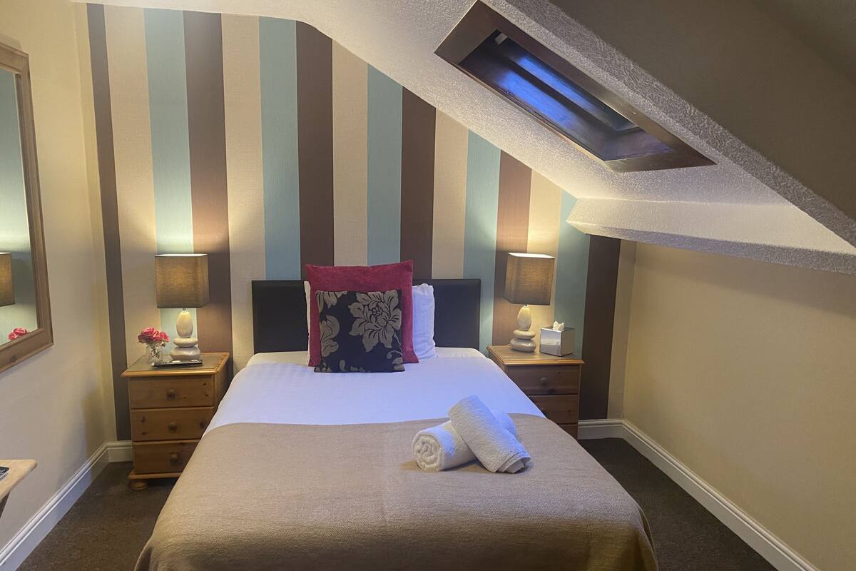 A neatly made bed with a striped wall behind it, a bedside table with a lamp, and a small vase with flowers.