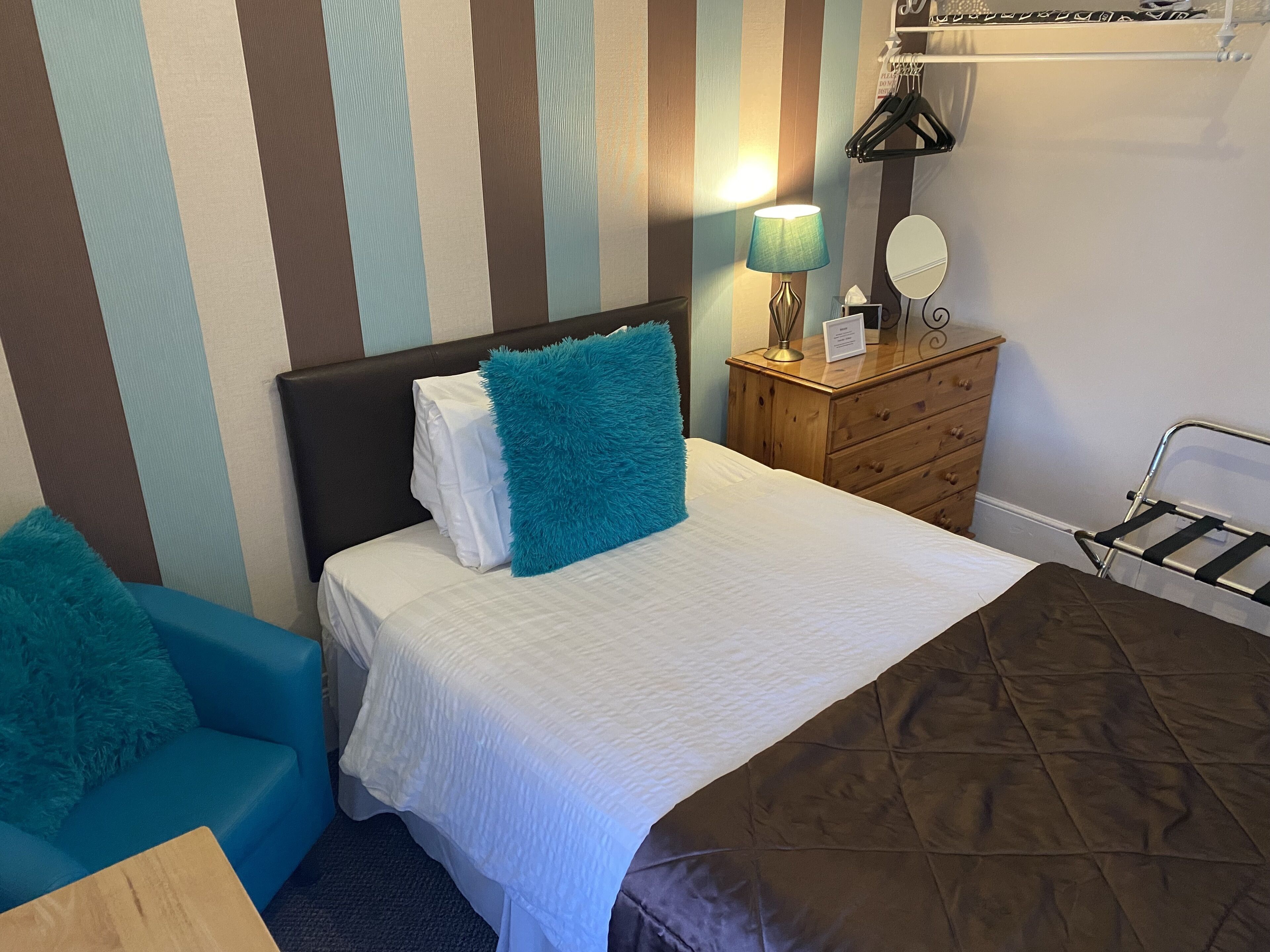 A hotel room with a bed, a wooden dresser, a lamp, and a striped wall.