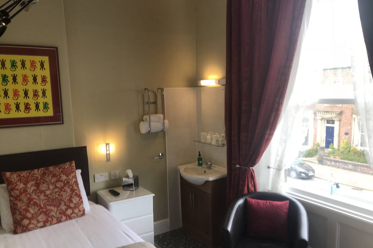 A hotel room with a bed, a bedside table with a lamp, a chair, a bathroom with a sink and mirror, and a window with a view of the street.