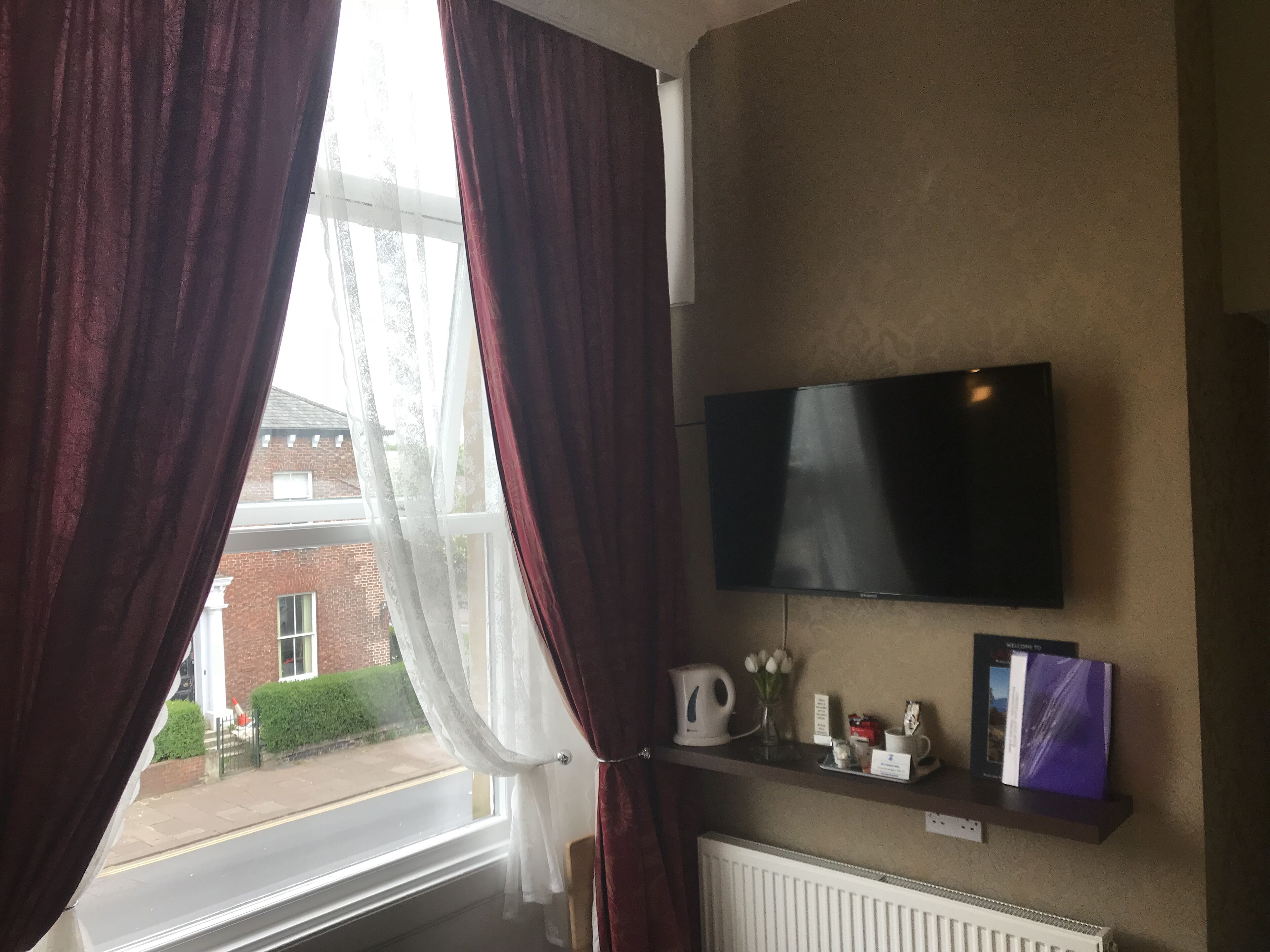 A room with a flat-screen TV mounted on the wall, a radiator below it, and a shelf with various items including a kettle, a vase with flowers, and a book.
