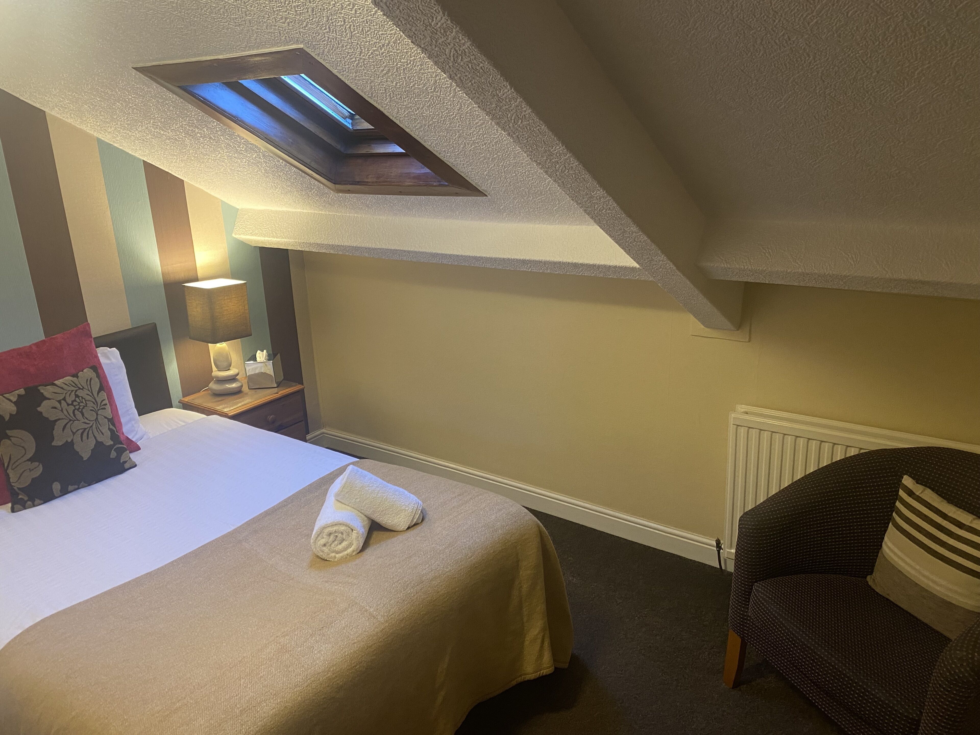 A hotel room with a bed, a nightstand, a lamp, a chair, and a skylight.