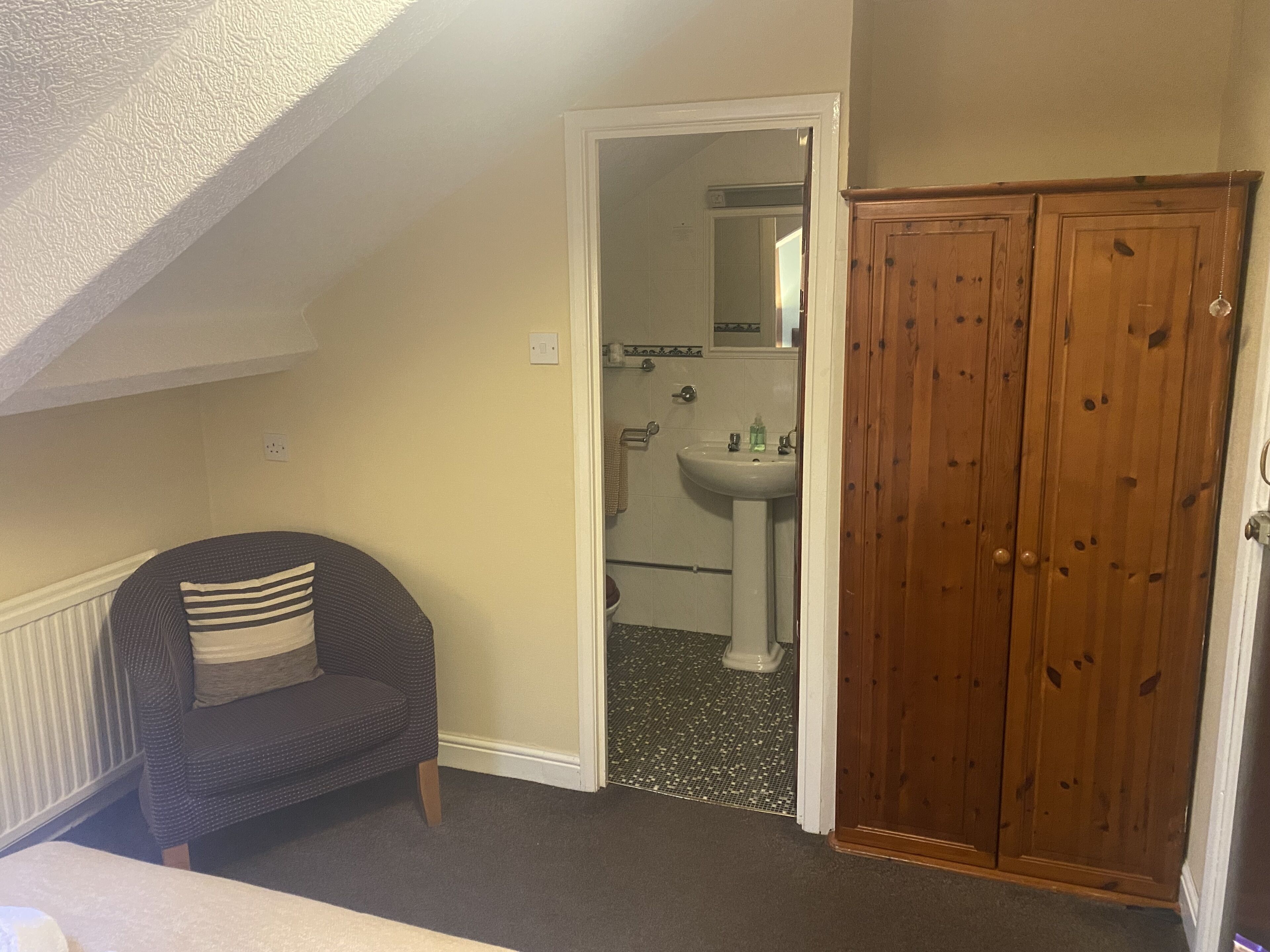 Une pièce avec un plafond en pente, un fauteuil sombre agrémenté d’un coussin à rayures, une armoire en bois et une salle de bain avec un lavabo et un miroir.