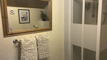 A bathroom with a wooden-framed mirror, two fluffy towels, a toilet paper holder, and a glass-enclosed shower.