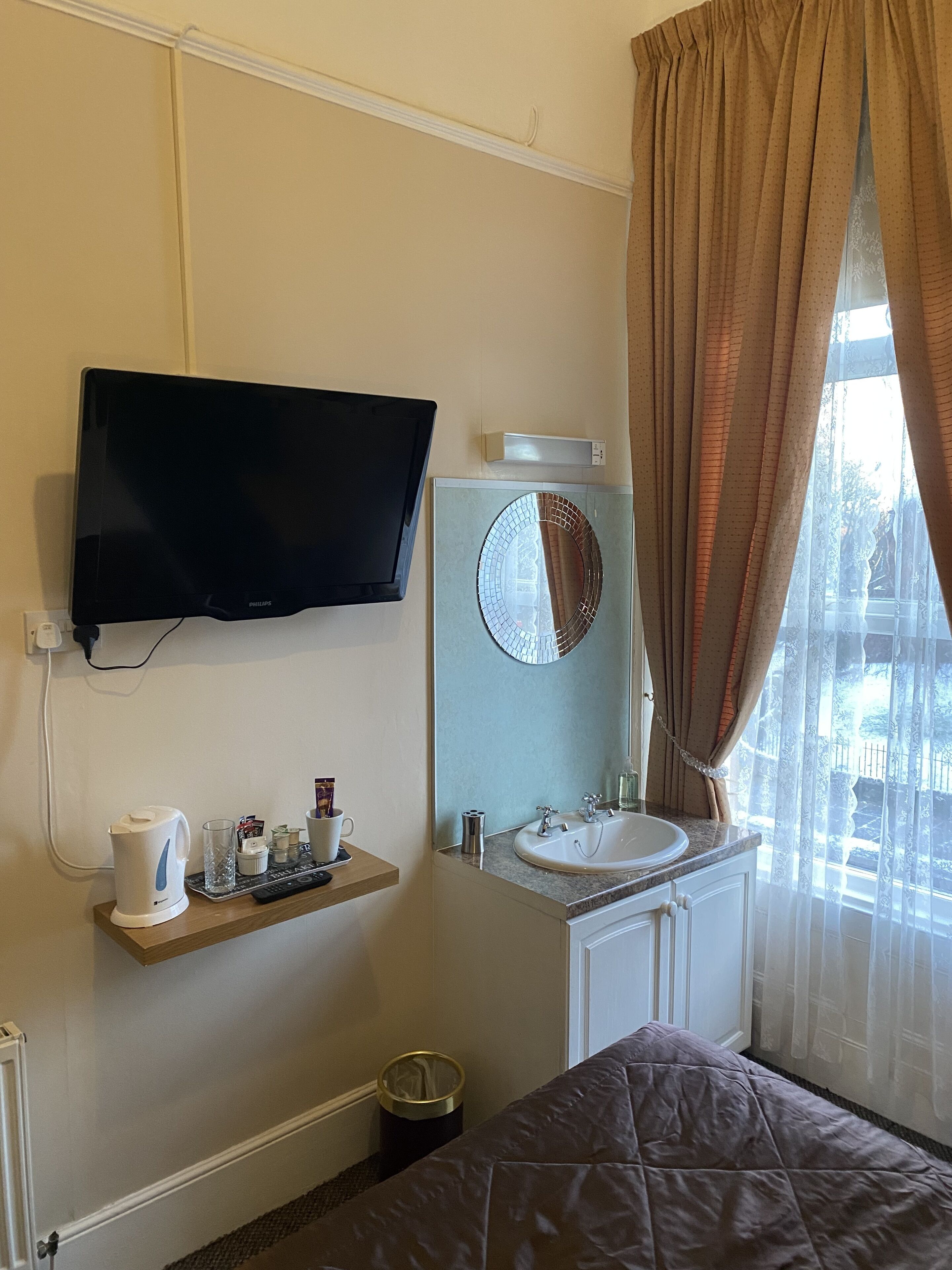 A hotel room with a television mounted on the wall, a sink with a mirror, a bed with a brown blanket, and a window with curtains.