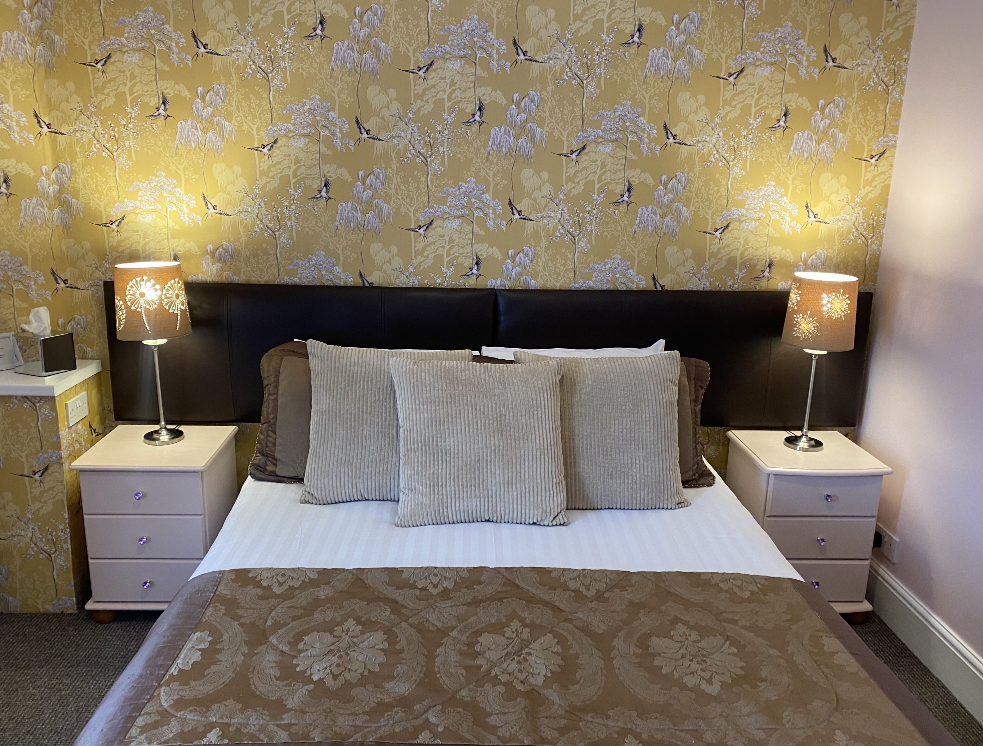 A neatly made bed with a floral-patterned bedspread, two nightstands with lamps, and a wall with a bird and tree wallpaper.