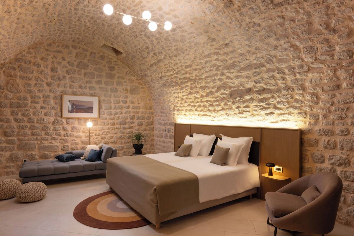 Une chambre confortable avec un mur en pierre, un lit doté d’une tête de lit en bois, une table de chevet avec une lampe, un canapé et une plante en pot.