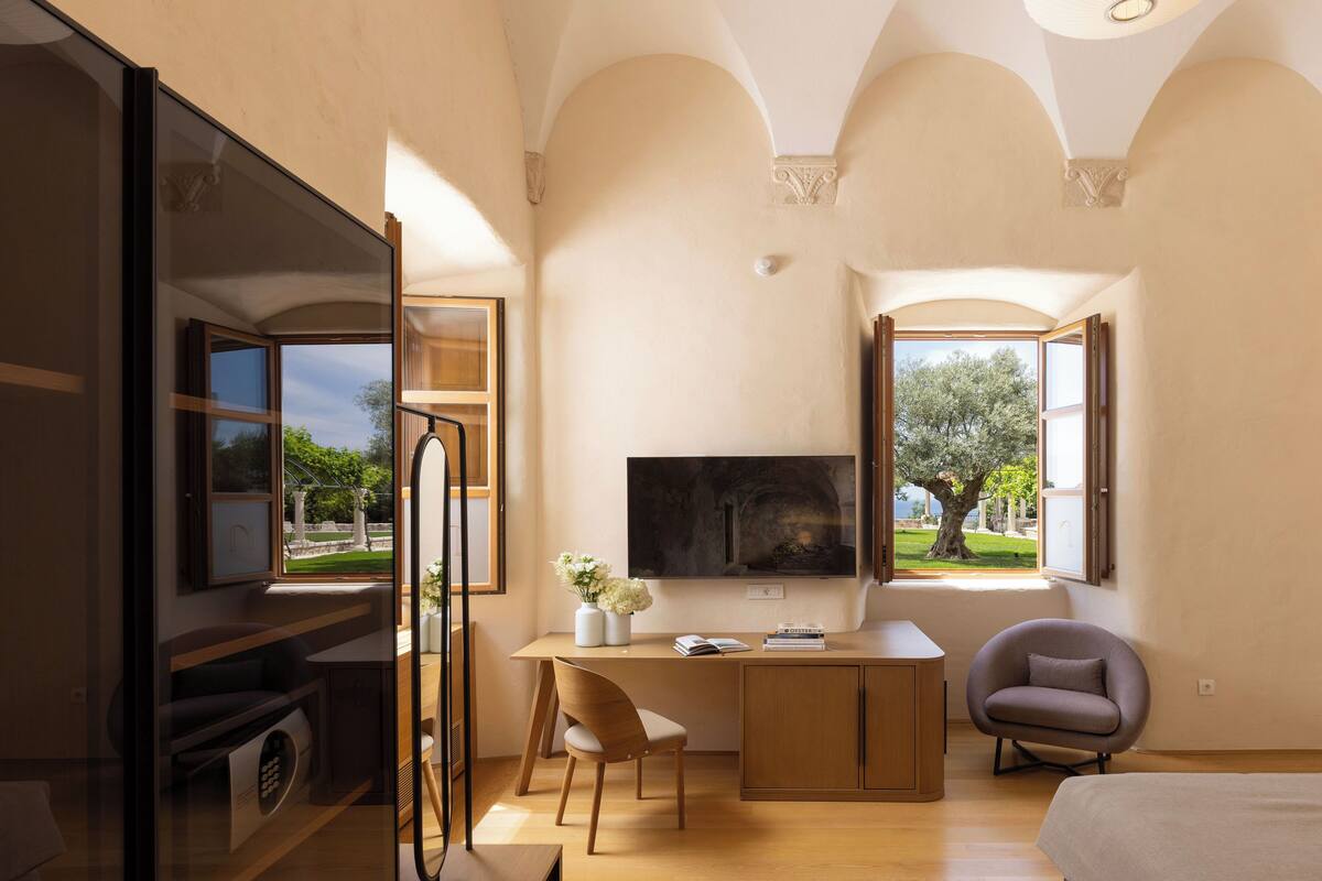 Une chambre moderne avec un grand lit, un bureau avec une chaise, une télévision et deux fenêtres donnant sur l’extérieur.
