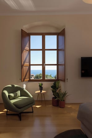 Una habitación con un ventanal grande, un sillón verde, una mesita y dos plantas en macetas.