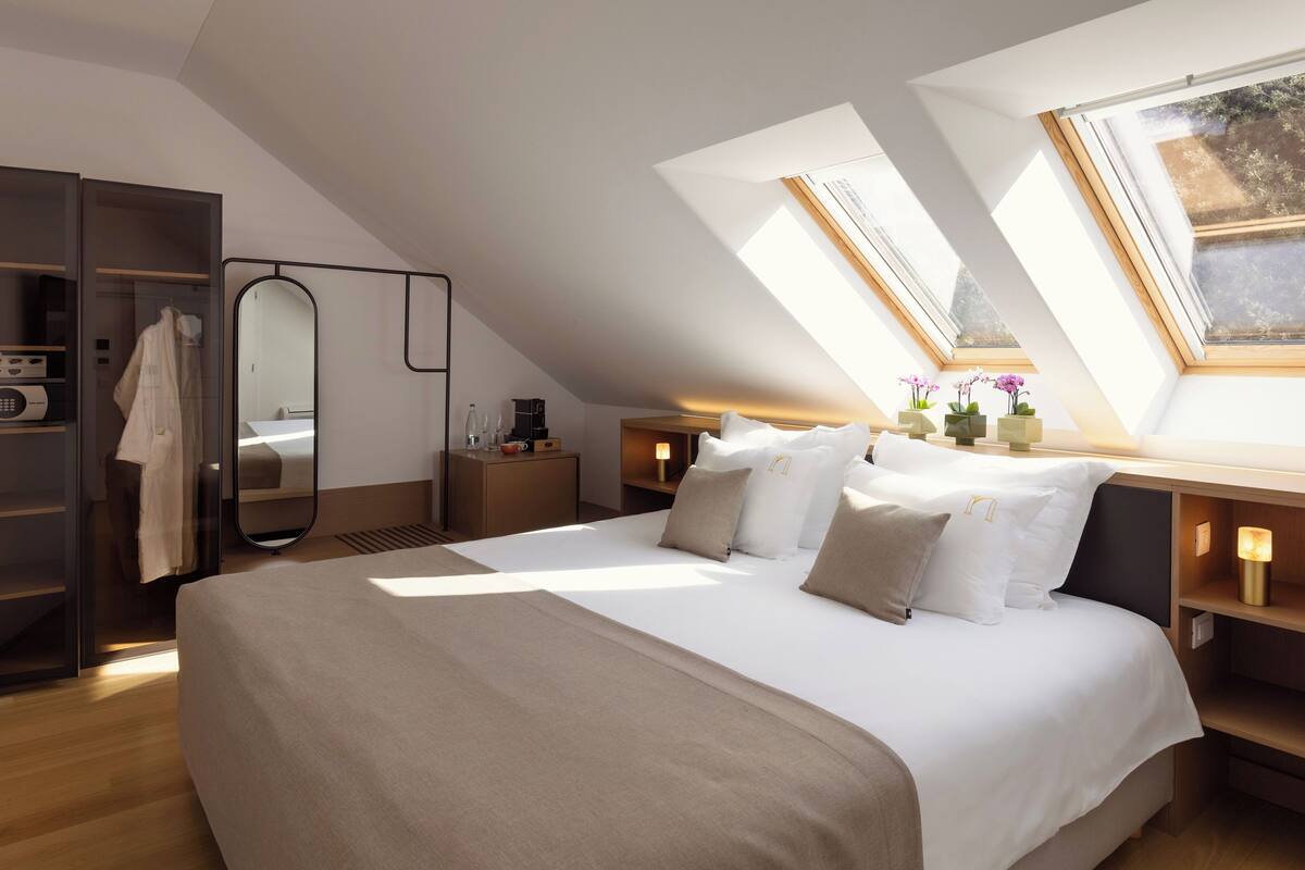 Quarto moderno com claraboia, cama com roupa de cama branca, mesa de cabeceira com abajur e guarda-roupa.
