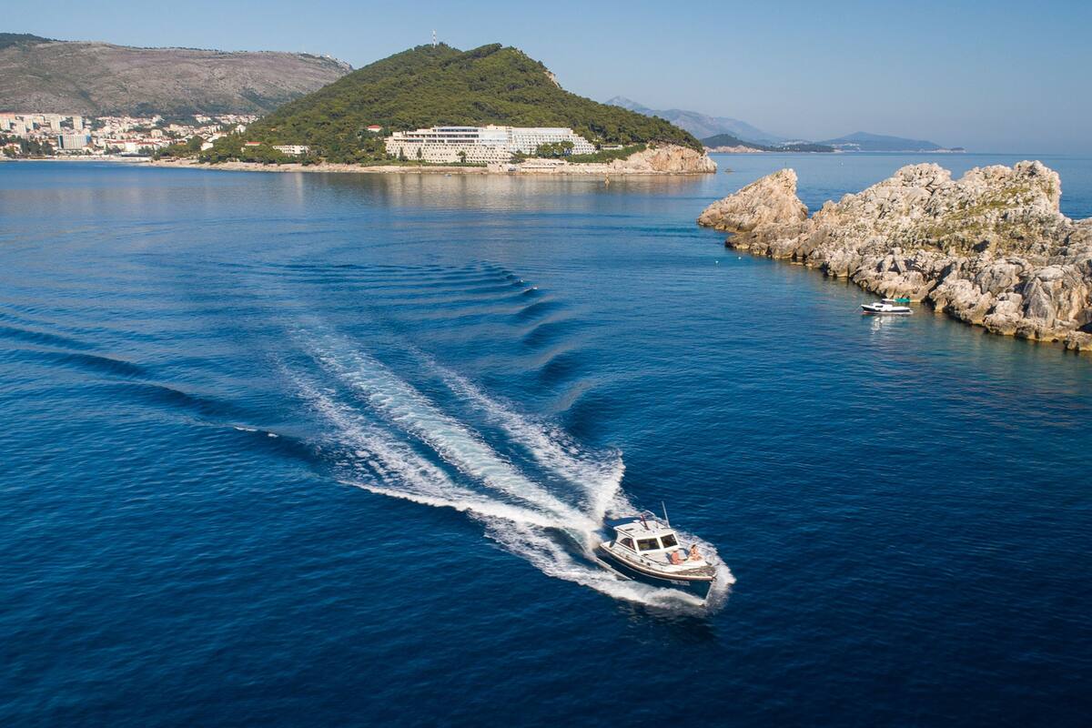 Un bateau qui laisse une vague derrière lui sur une étendue d’eau importante, avec une côte rocheuse et une colline parsemée de bâtiments en arrière-plan.