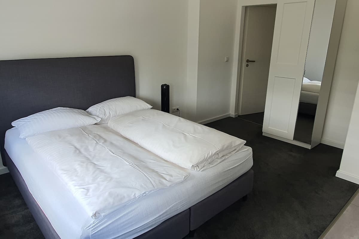 A neatly made bed with white linens in a room with a dark floor and a grey headboard.