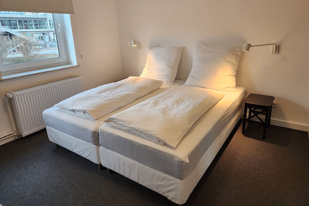 A double bed with white bedding, a small bedside table, a lamp, and a view of a building through a window.