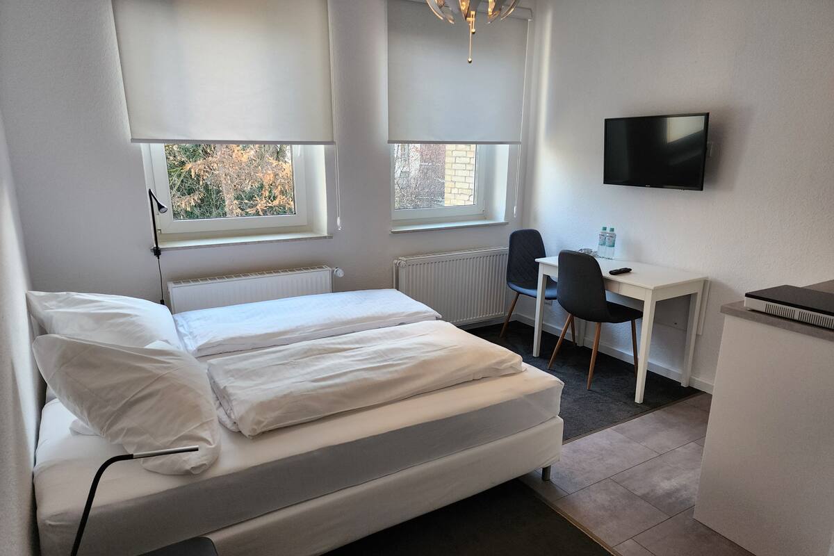 A hotel room with a bed, a desk, a chair, a TV, and windows with blinds.