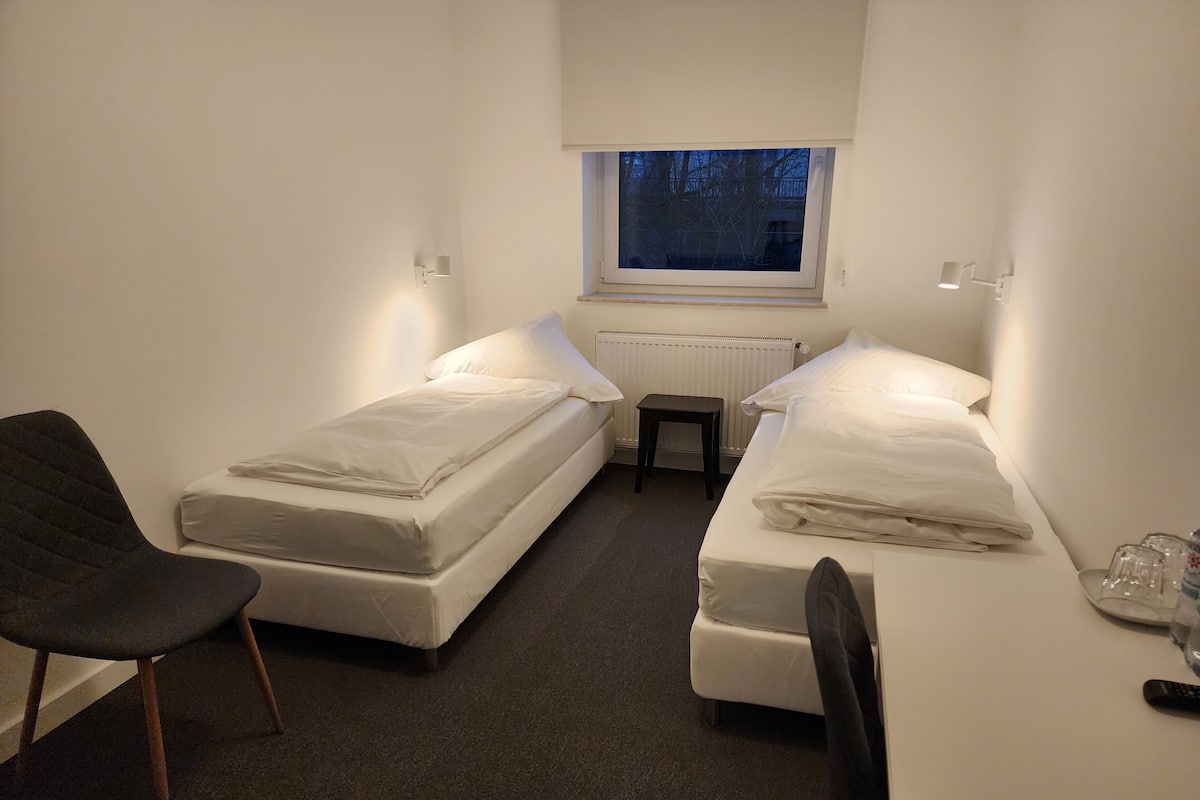 A small, compact room with two single beds, a chair, a small table, and a window with a view of trees.