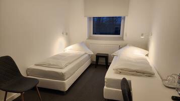 Ein kleiner, kompakter Raum mit zwei Einzelbetten, einem Stuhl, einem kleinen Tisch und einem Fenster mit Blick auf Bäume.
