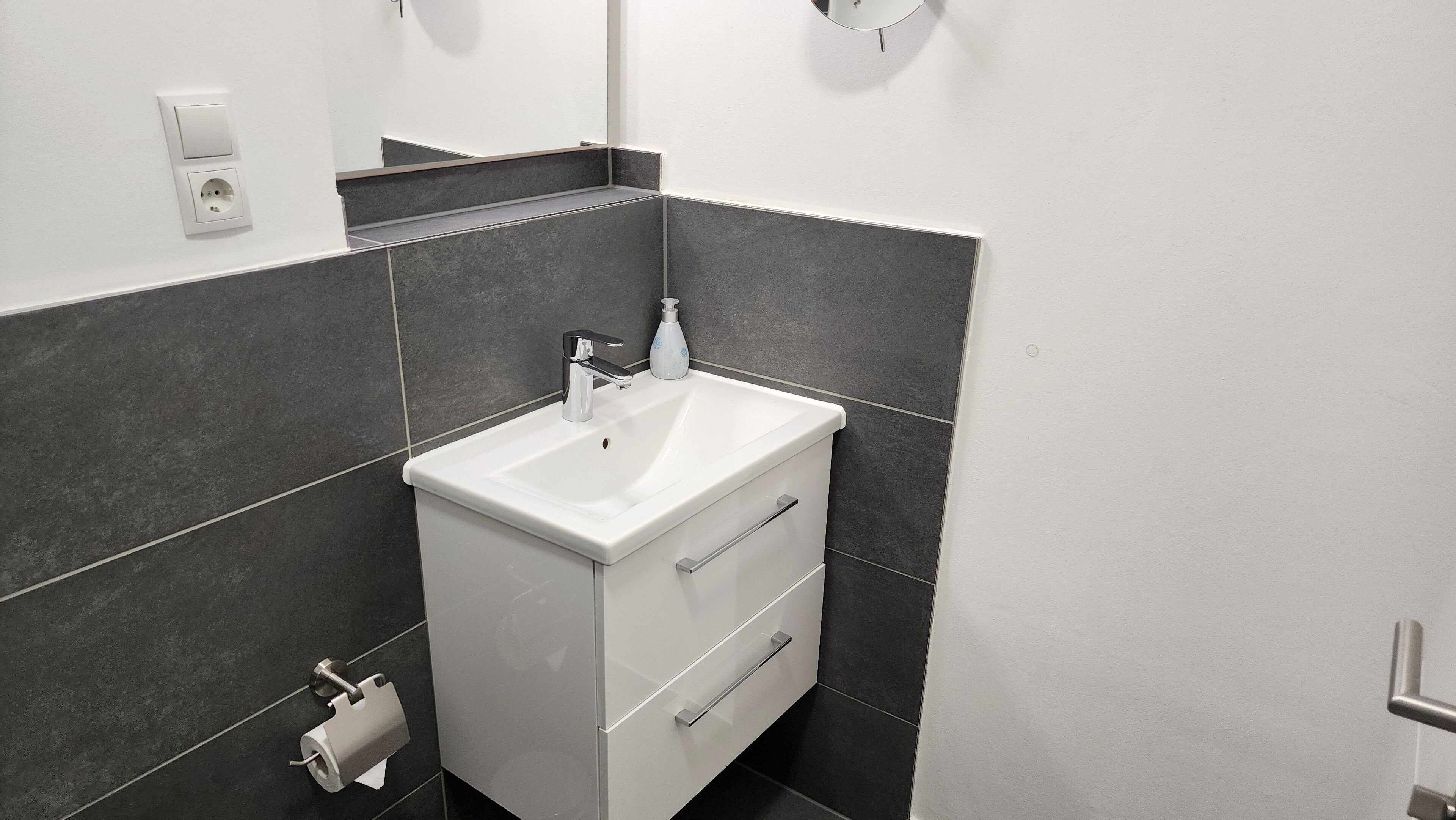 A bathroom with a white sink, dark tiled walls, a mirror, a soap dispenser, and a toilet paper holder.