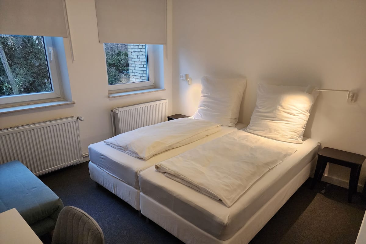 A single-bed room with a window, a radiator, a bedside table, and a chair.