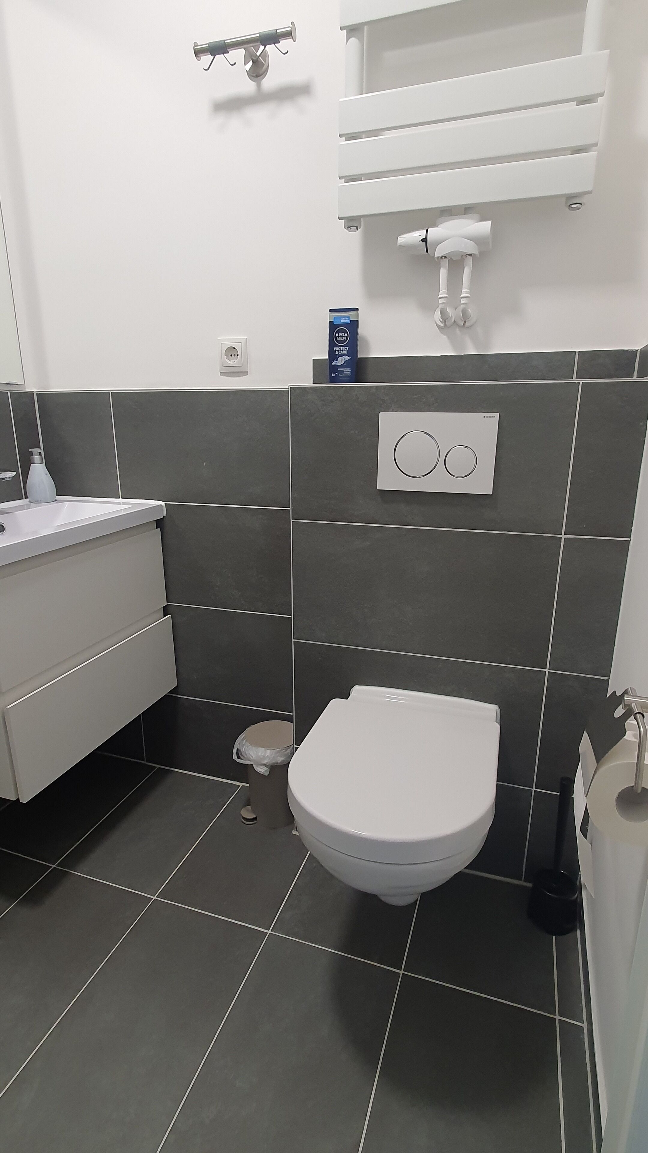 Ein modernes Badezimmer mit Toilette in Weiß, grauen Fliesen, einem weißen Waschbecken und einer kleinen Mülleimer.