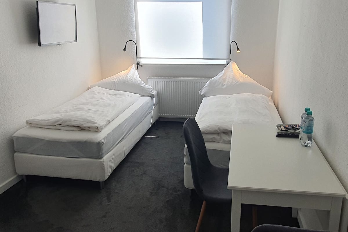 A small, compact room with two single beds, a small table, and a chair. There is a window providing natural light and a radiator below the window.