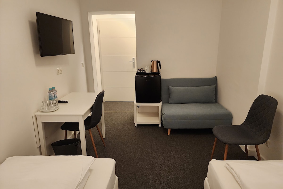 A hotel room with a bed, a desk with a chair, a sofa, a TV, and a kitchenette.
