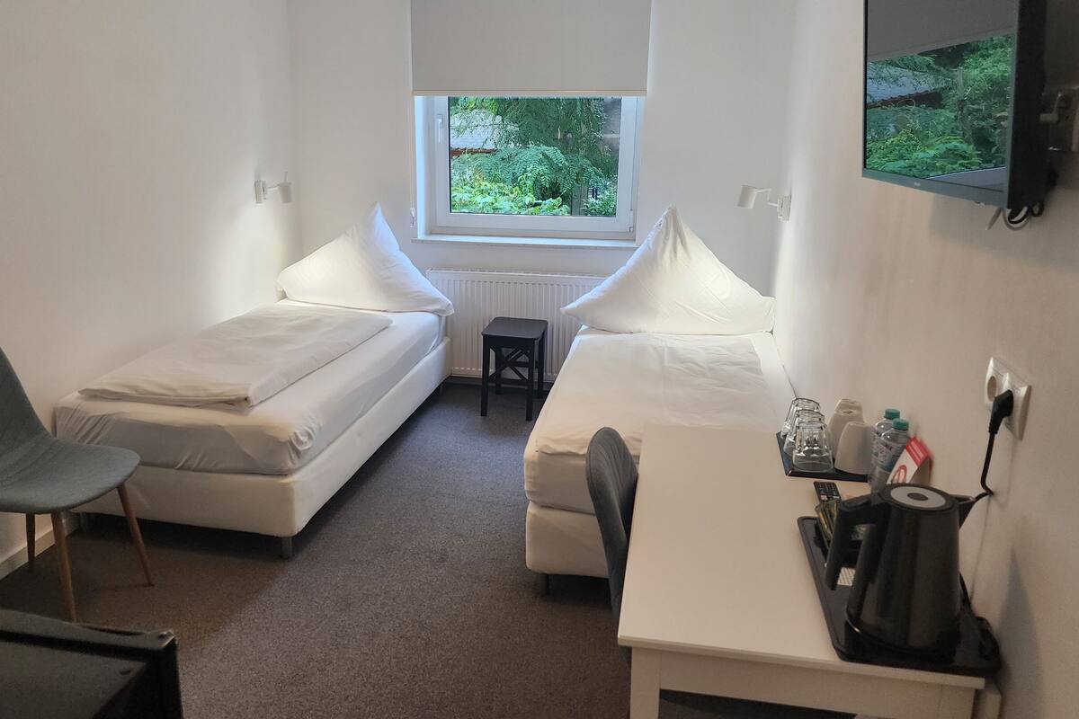 A small, clean room with two single beds, a desk with a chair, a small table, and a window with a view of greenery.