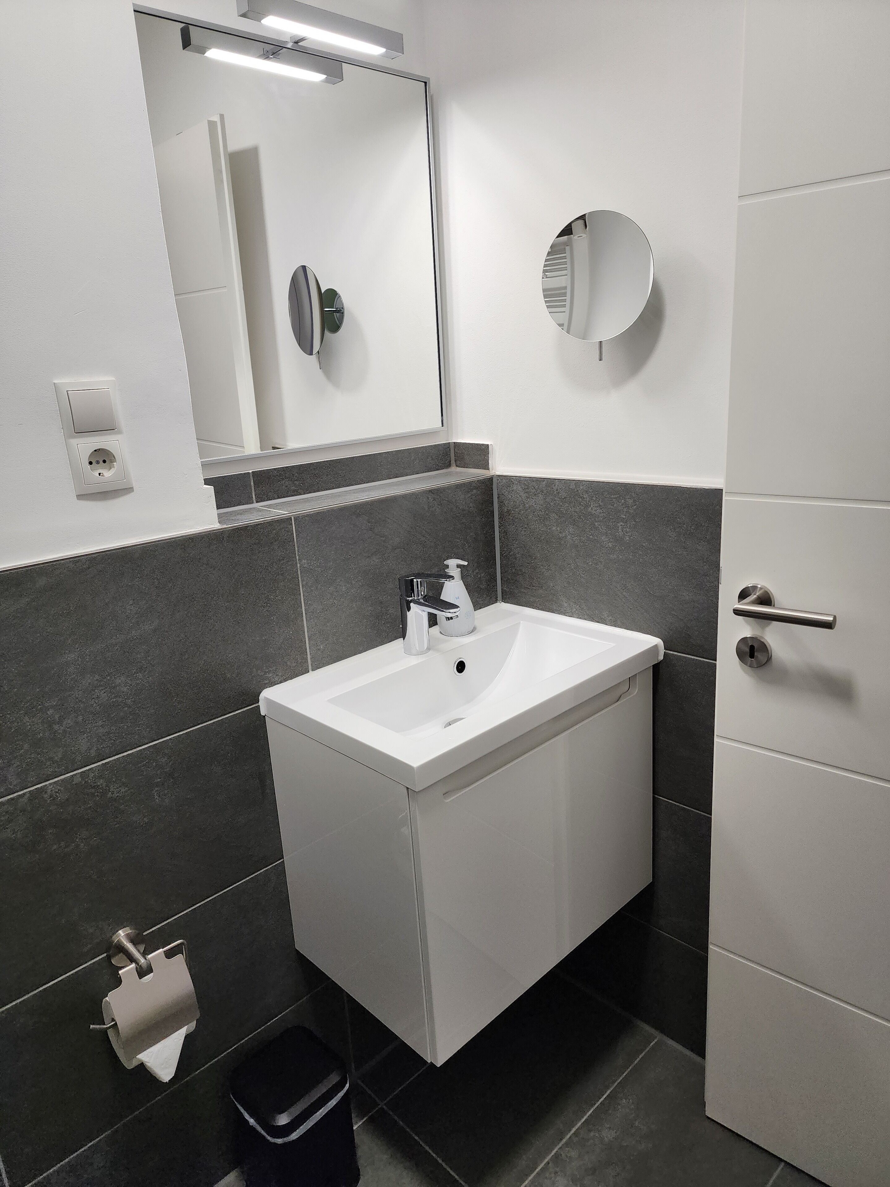 Een moderne badkamer met een witte wastafel, donkere betegelde wanden, een spiegel en een toiletrolhouder.