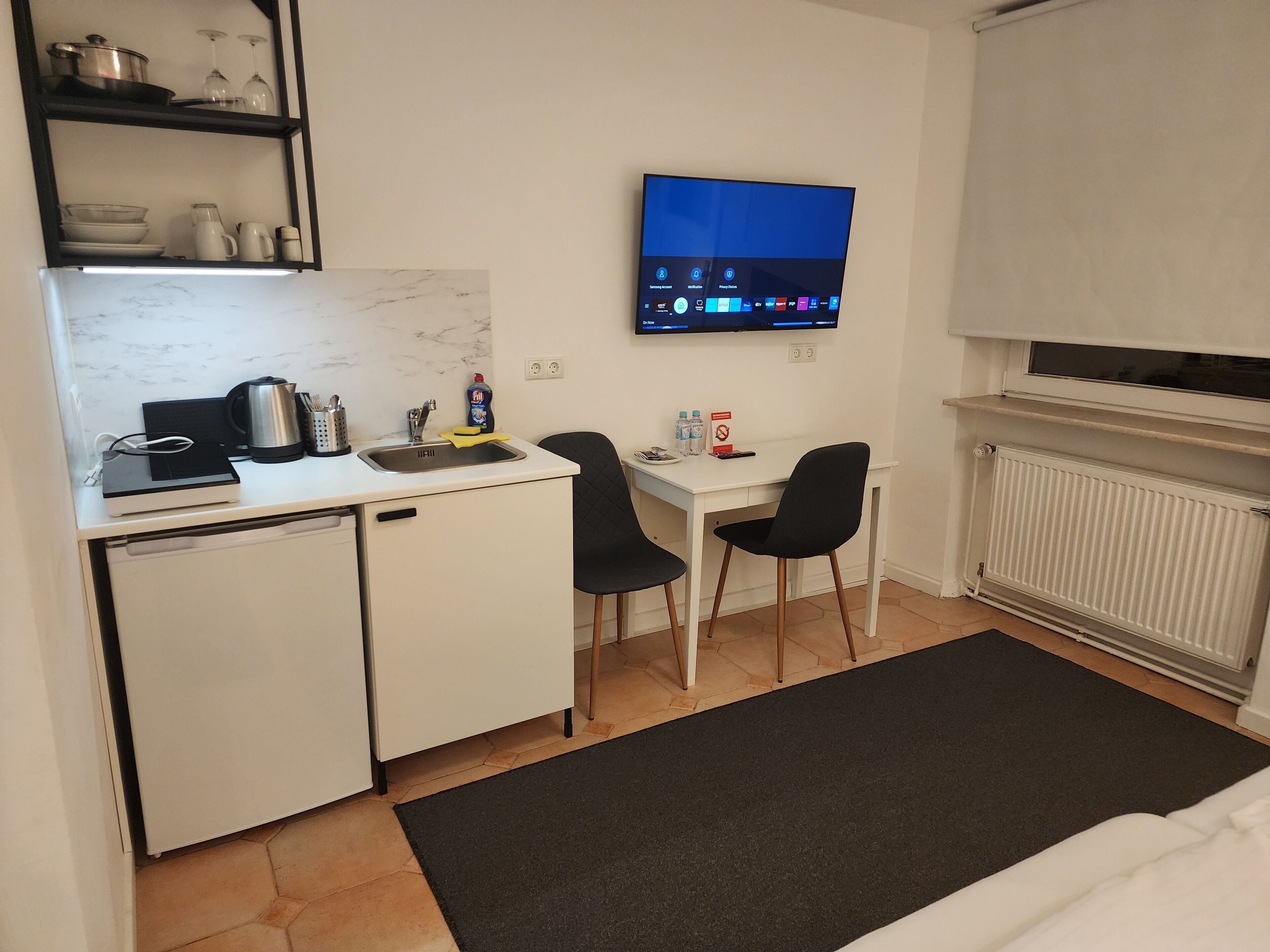 Une petite cuisine équipée avec un petit réfrigérateur, un évier et un four à micro-ondes. On y trouve une chaise noire et une petite table sur laquelle sont posés un ordinateur portable et une bouteille d’eau. Un téléviseur à écran plat est fixé au mur, au-dessus de la table.