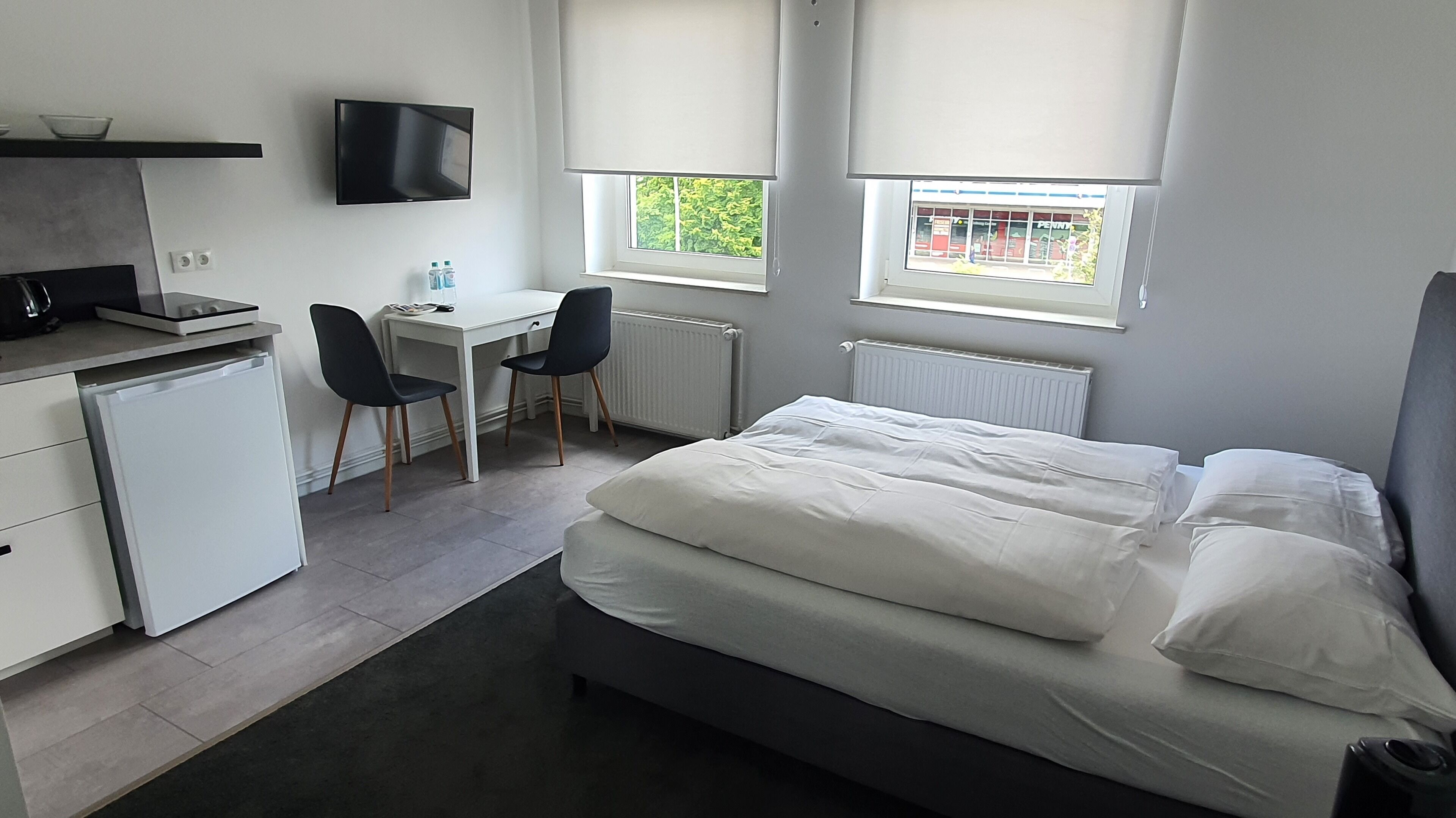 A compact hotel room with a bed, a desk, a chair, a small fridge, and a TV.