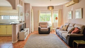 Ein Wohnzimmer mit Sofa, Sessel und Teppich. Es gibt eine Küchenzeile mit Spüle und Schränken.