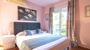 Ein Schlafzimmer mit einem großen Bett, einem Nachttisch, einer Wand-Klimaanlage, einem Fenster mit Vorhängen und Blick auf einen Balkon.
