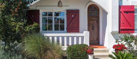 A house with a white exterior, a wooden porch, a red door, and a window with shutters.