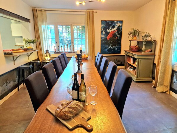 Une longue table à manger entourée de chaises, une planche à découper en bois avec une miche de pain, des bouteilles de vin et des verres.