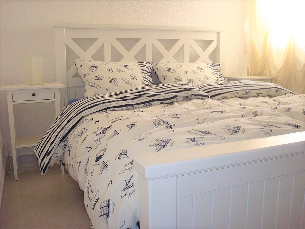 A neatly made bed with striped bedding and patterned pillows, a white nightstand with a lamp, and a white headboard with a geometric design.