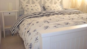 Ein ordentlich bezogenes Bett mit gestreifter Bettwäsche und gemusterten Kissen, ein weißer Nachttisch mit Lampe sowie ein weißer Kopfteil mit geometrischem Muster.