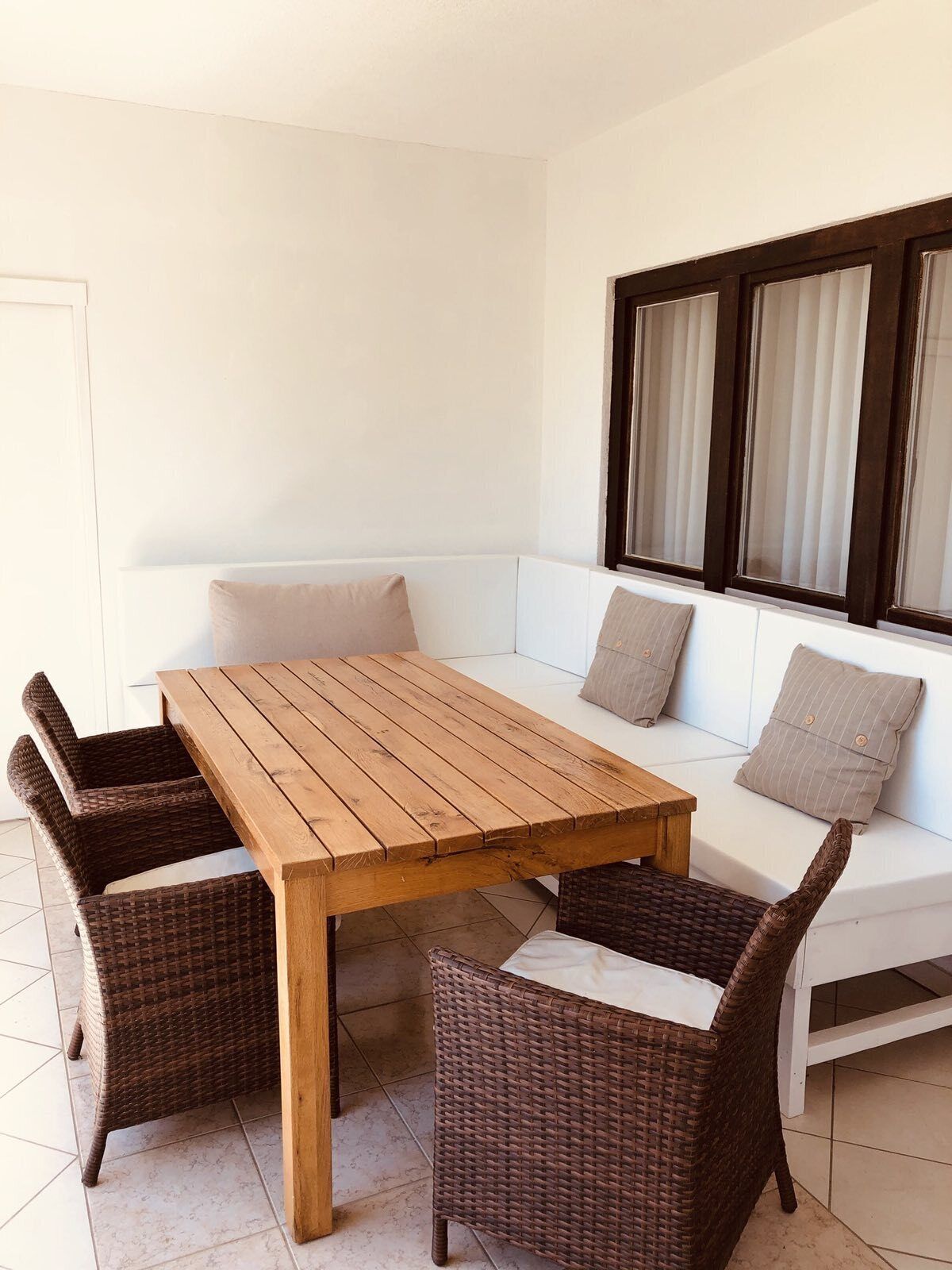 A wooden table with four chairs, a white sofa with cushions, and a window with blinds.