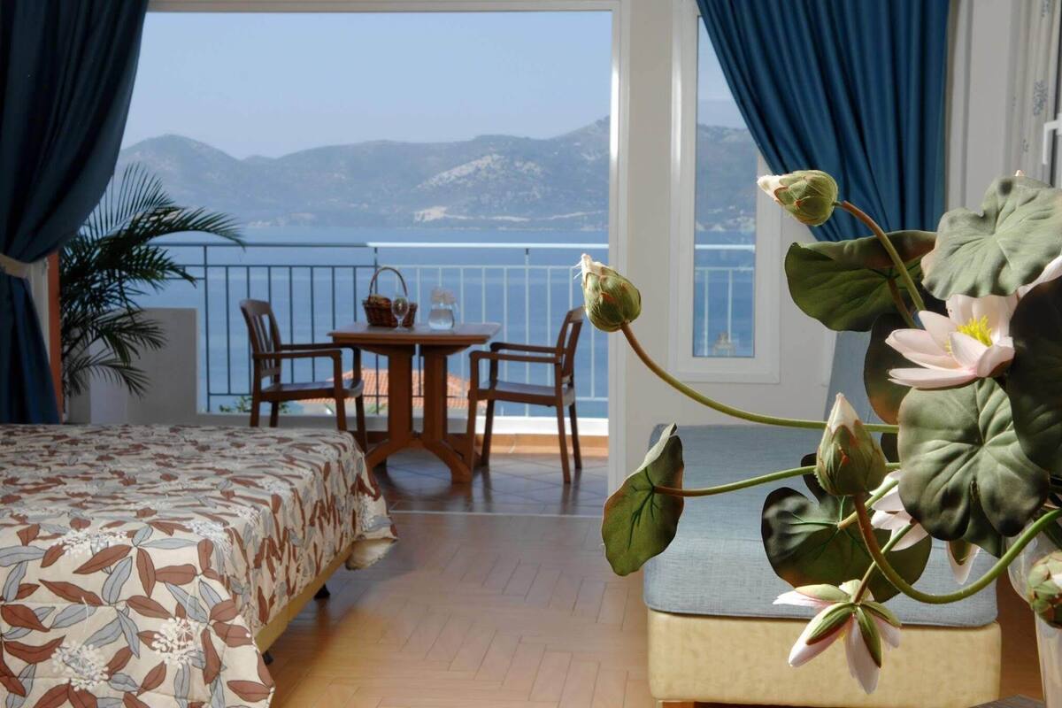 A bedroom with a bed, a table with chairs, a potted plant, and a view of the sea and mountains.