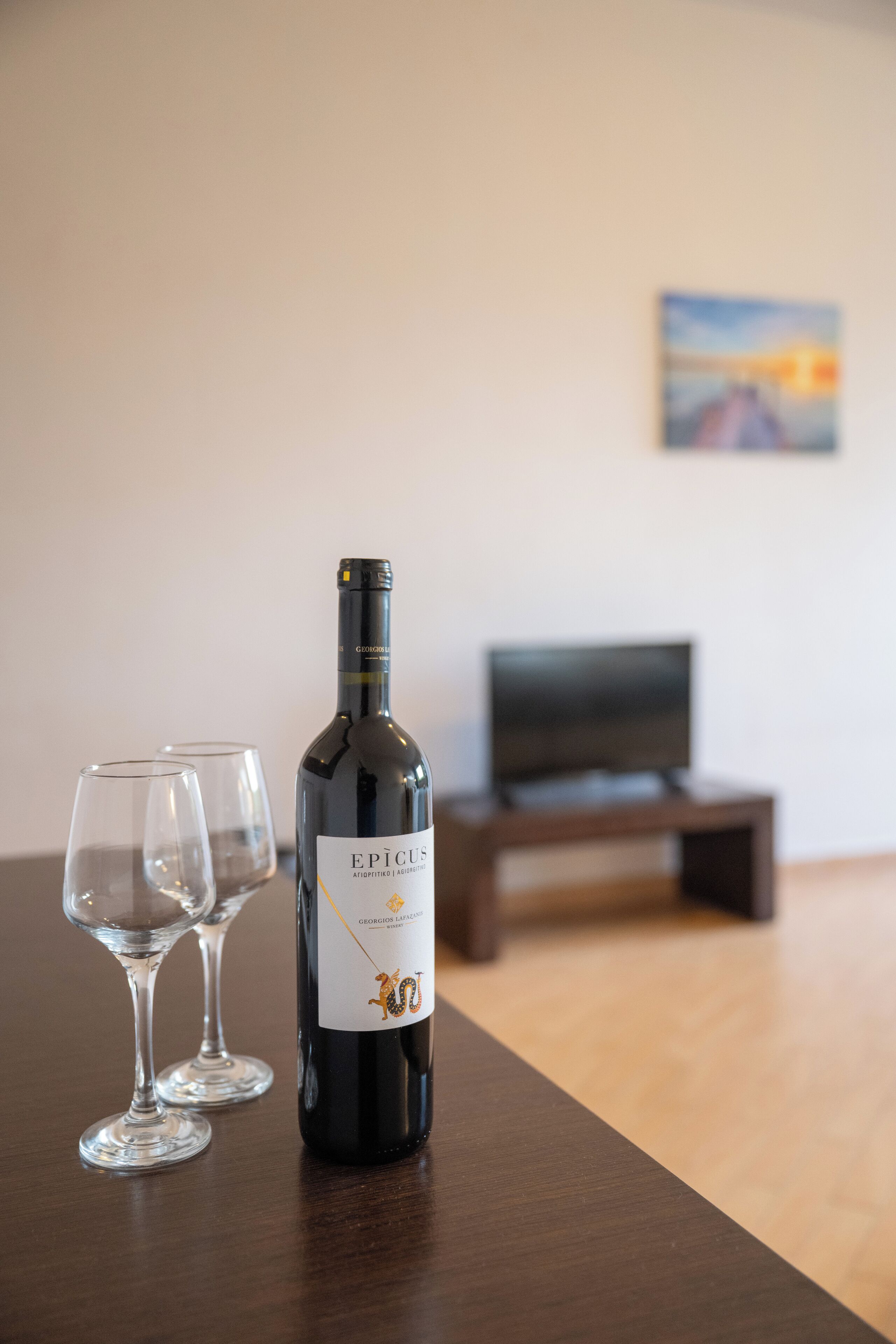 A bottle of wine and two glasses on a wooden table in a room with a television and a painting on the wall.