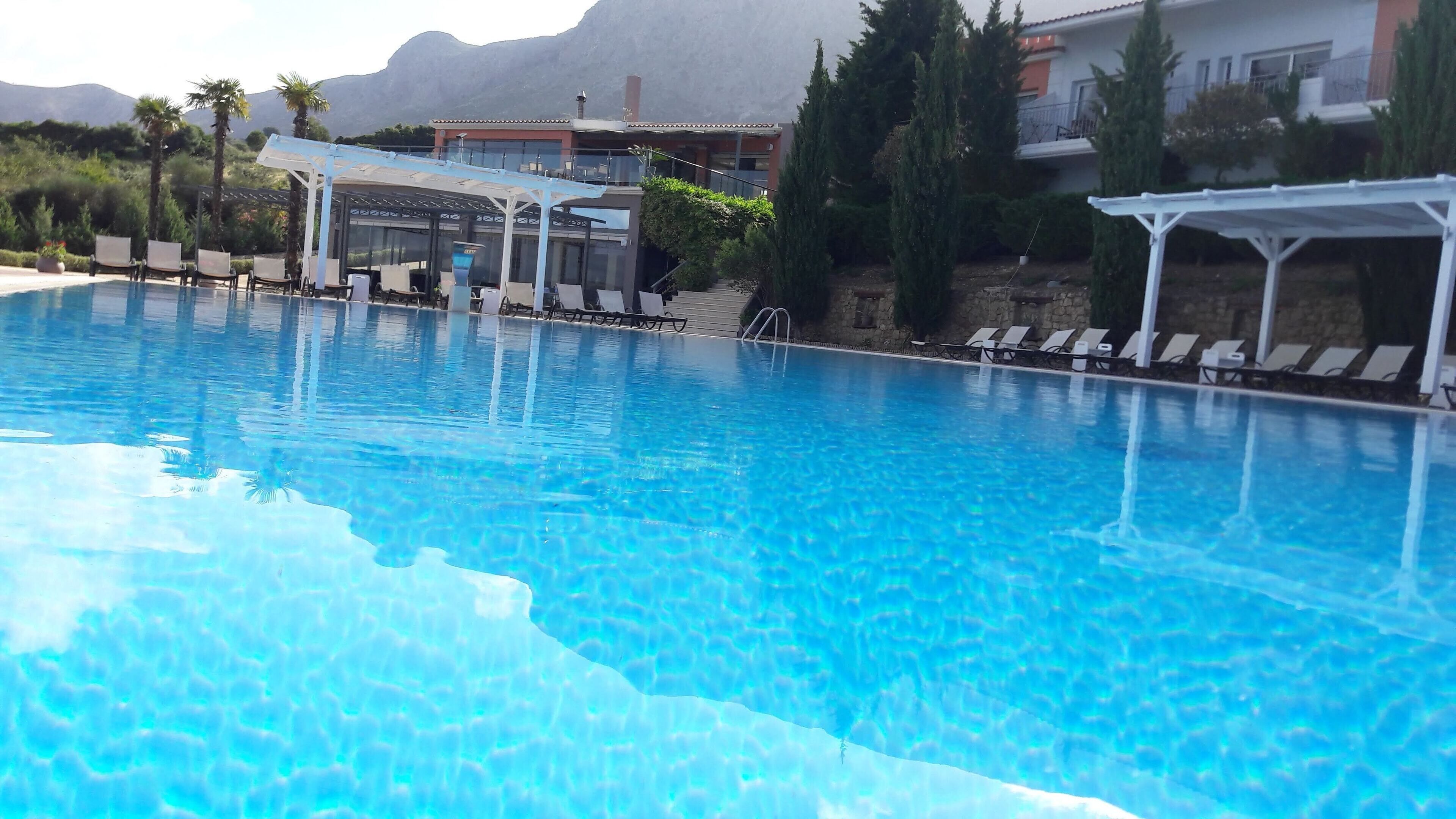 Piscina azul cristalina con sillas de descanso y una zona cubierta, rodeada de vegetación y edificios.