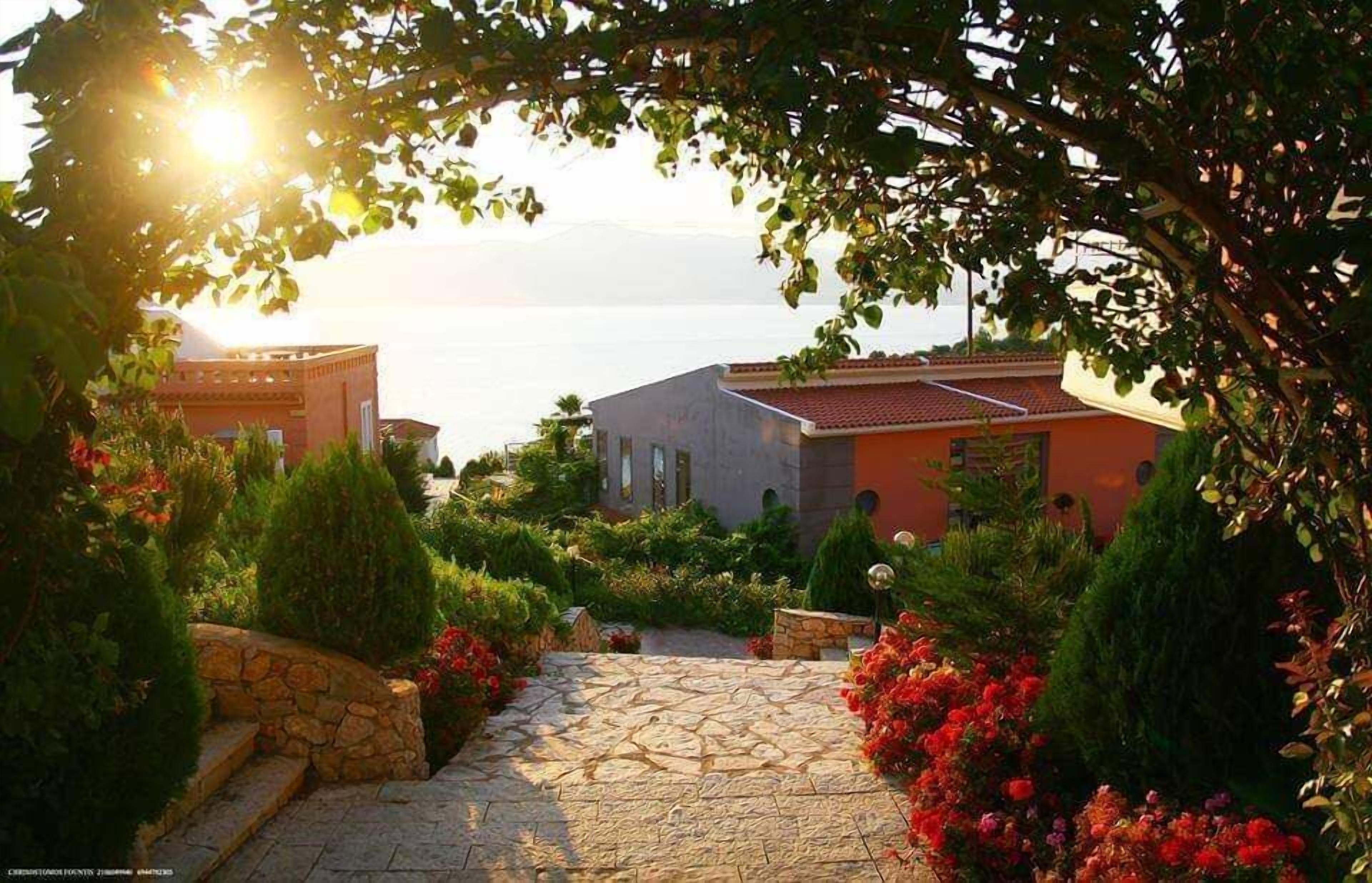 Un chemin de pierres mène à un bâtiment surmonté d’un toit rouge, qui est entouré d’une végétation luxuriante et de plantes en fleurs.