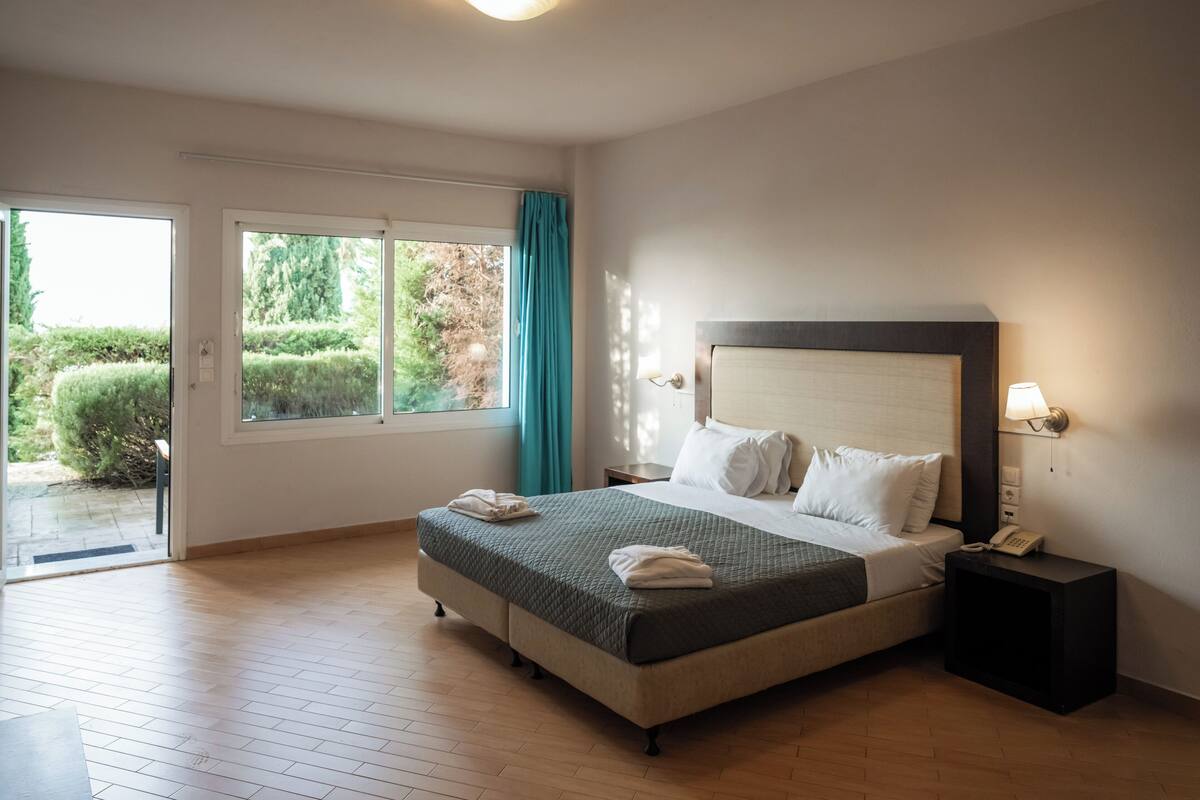 A hotel room with a bed, a nightstand, a lamp, and a view of greenery outside.