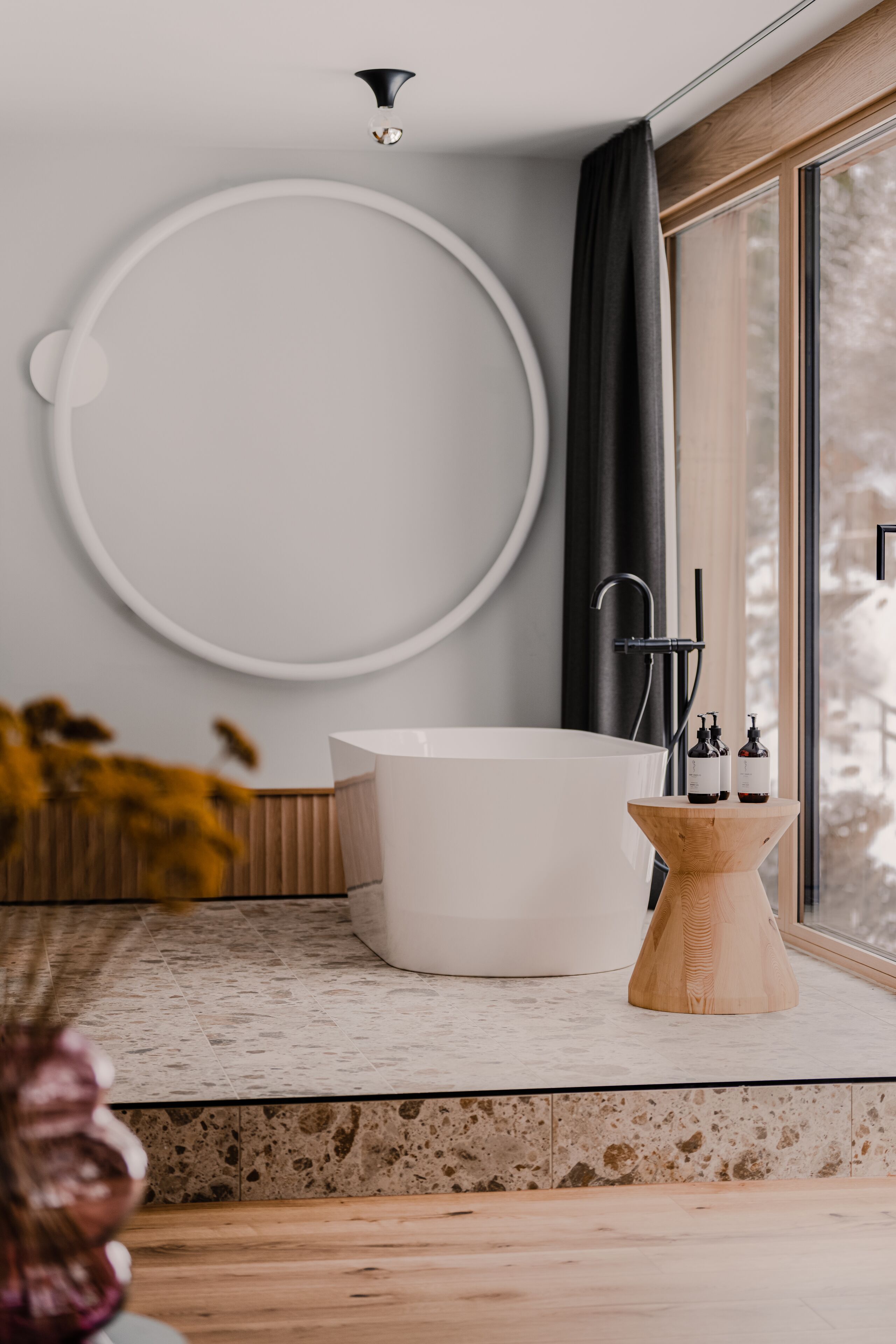 Un baño moderno con una bañera independiente, una mesita de madera con botellas y un espejo grande de forma circular en la pared.