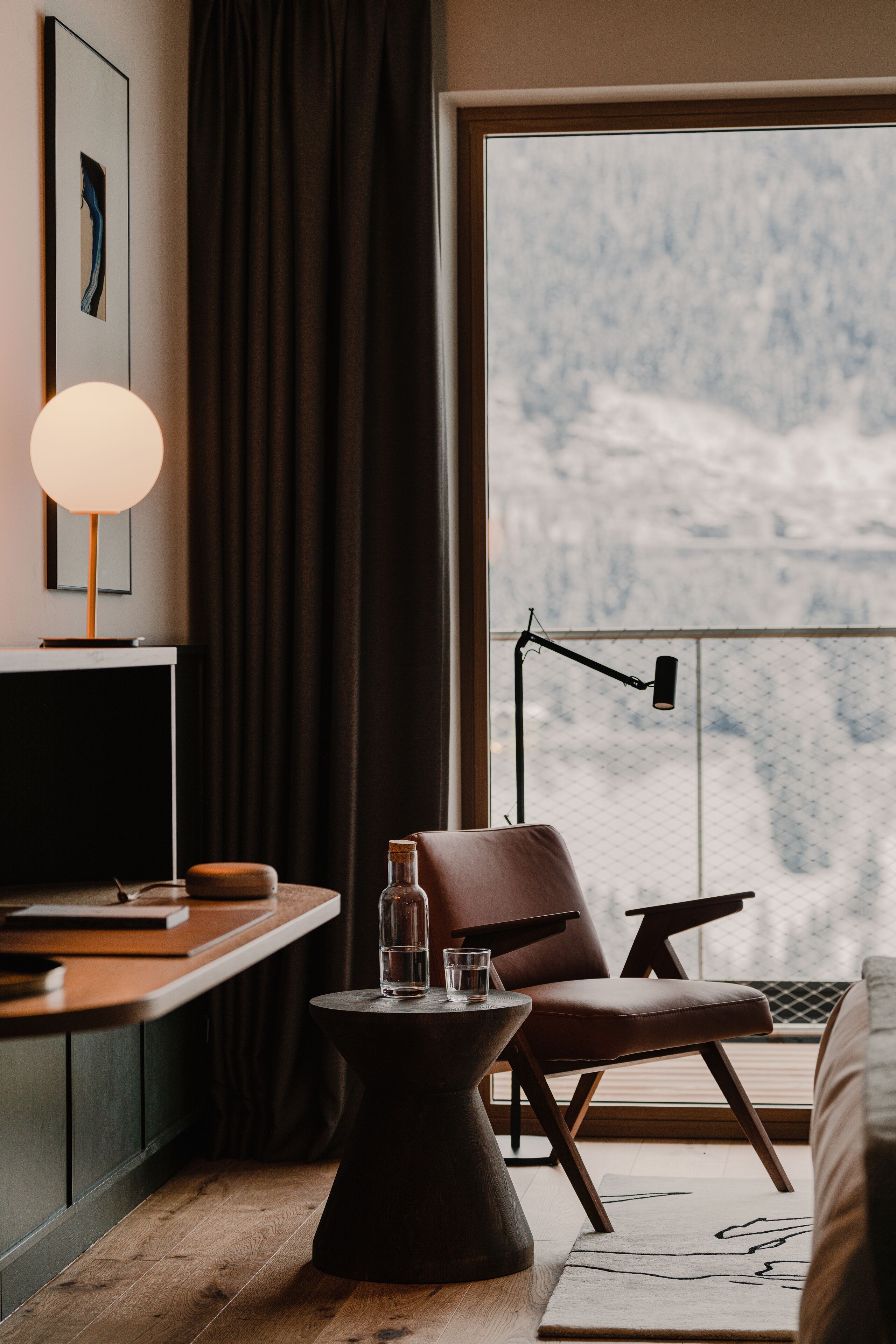 A modern hotel room with a large window, a desk with a lamp, a chair, and a small side table.